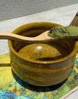 Matcha powder on wooden spoon with matcha bowl, bamboo whisk, and colorful mat