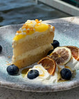 Slice of layered lemon cake with cream, topped with pearls, garnished with dehydrated citrus, blueberries, and whipped cream on a blue ceramic plate.