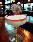 Frozen tropical cocktail garnished with dragon fruit, dried berries, and edible flower on bar.